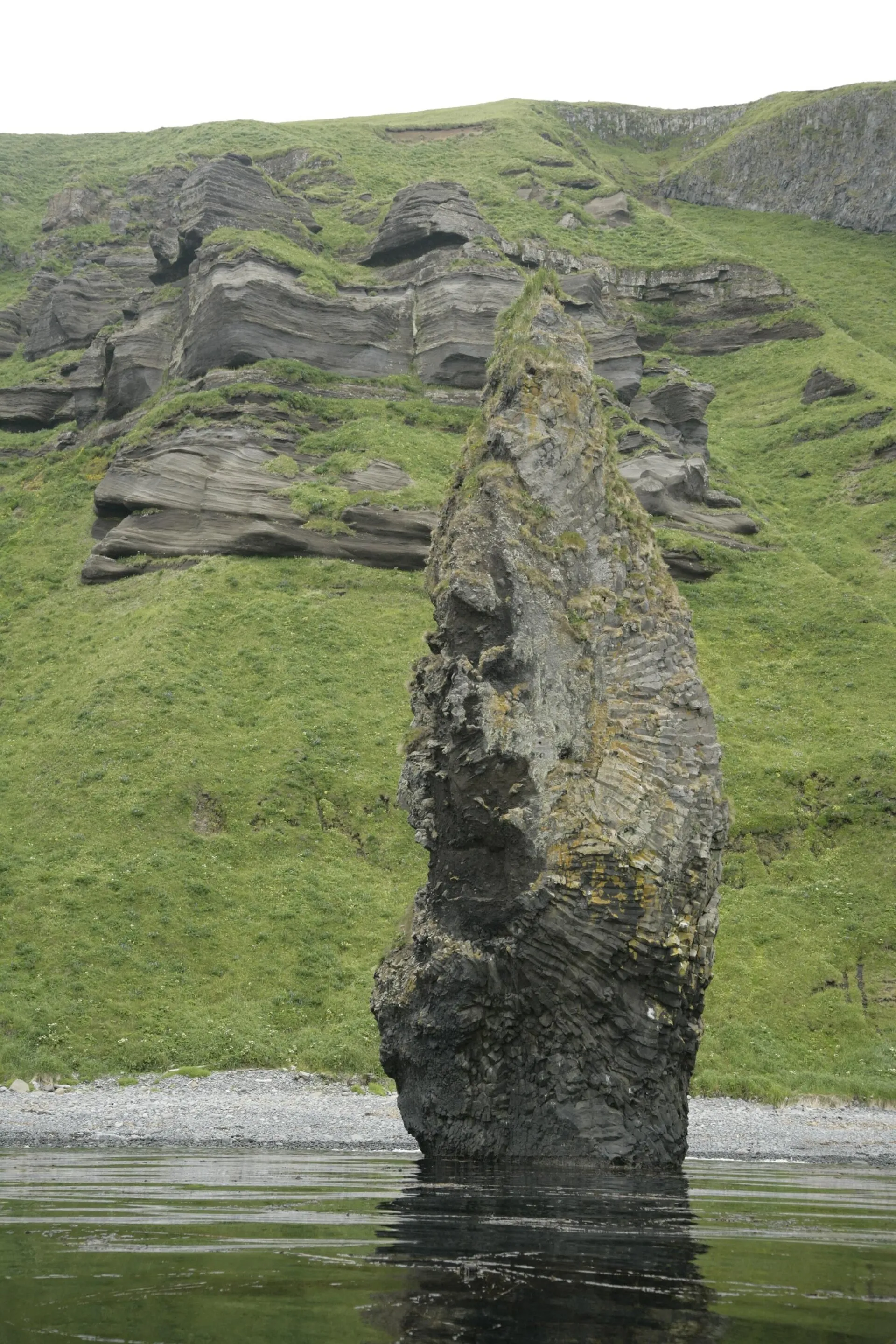 Basalt sea stack rising from the water beside green cliffs